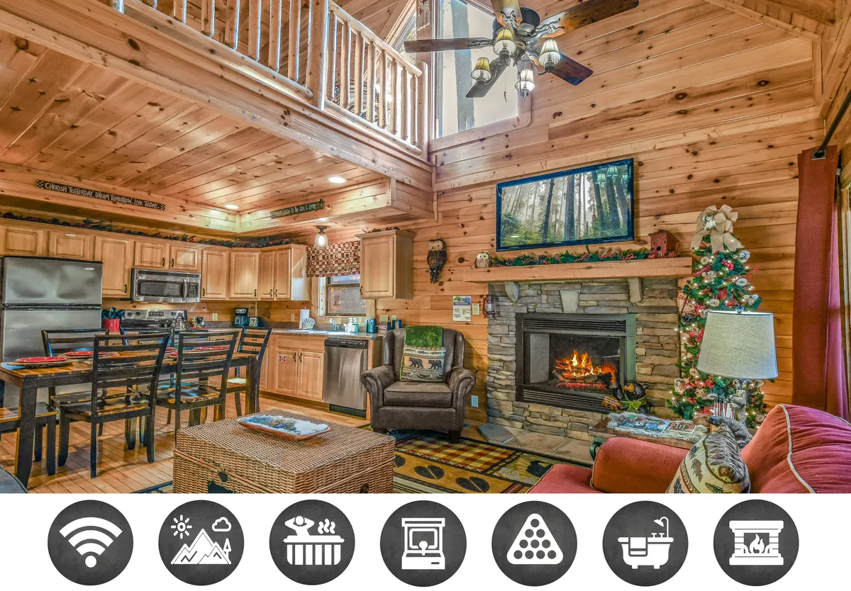 The image showcases a warm, inviting interior living space with a crackling fireplace as the focal point. An open-concept layout highlights the modern kitchen island perfect for cooking together, a cozy dining area set for shared meals, and a loft area above. Tasteful decorative artwork and mountain-themed accents add to the cabin's charm, creating a welcoming atmosphere for friends and families to gather, relax, and make memories.