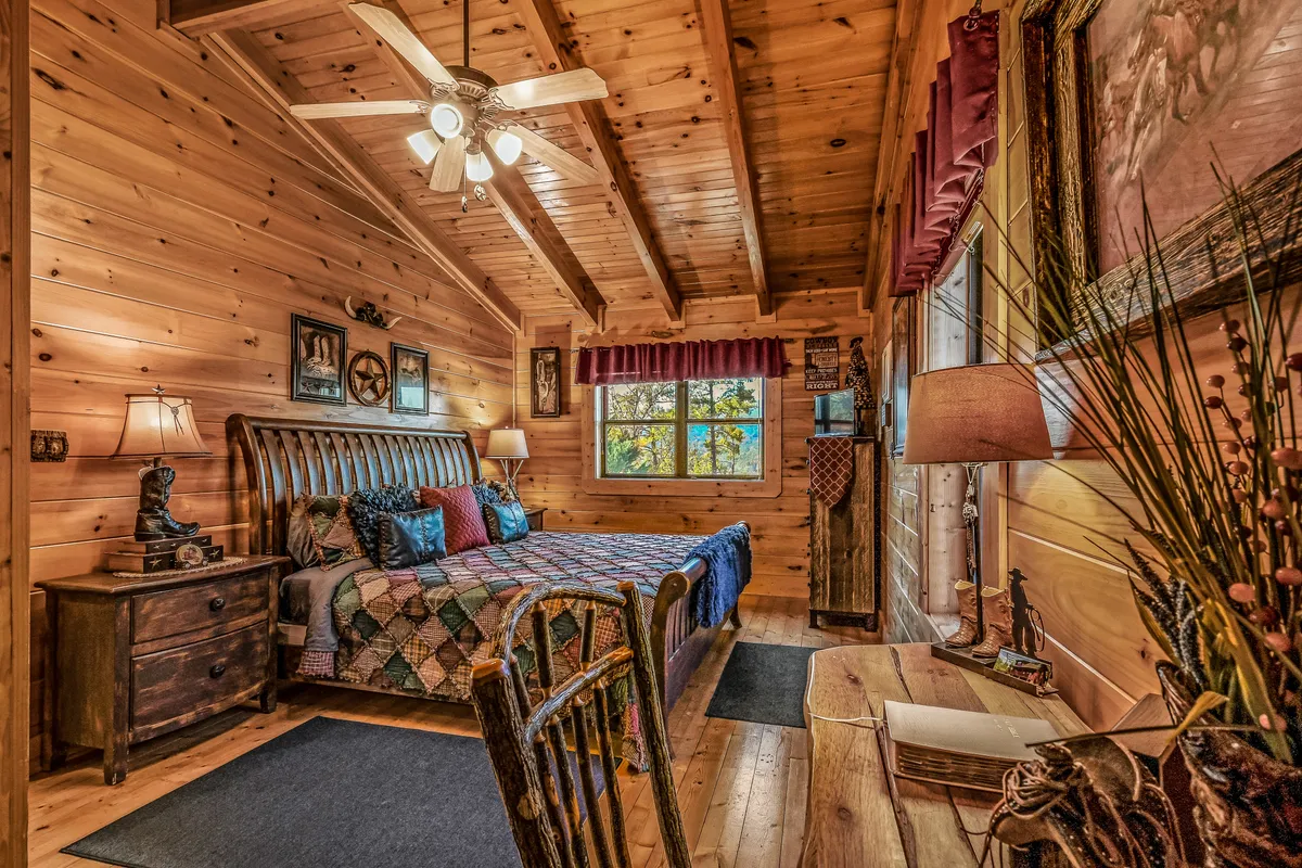 This room features a wooden bed with a colorful quilt, a window view bringing in natural light, artwork on the walls, and a wooden desk—subtly echoing the writing desk mentioned for the upstairs suite. The overall mood is welcoming and serene, ideal for quiet mornings or getting some work done.