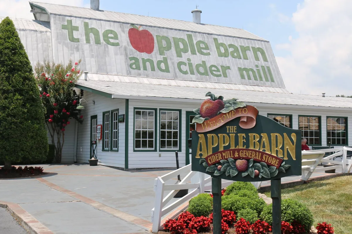 A welcoming entrance to a local attraction featuring a large apple-themed sign, flower beds, and inviting doors, suggesting opportunities for shopping, tasting local treats, and enjoying a friendly atmosphere.