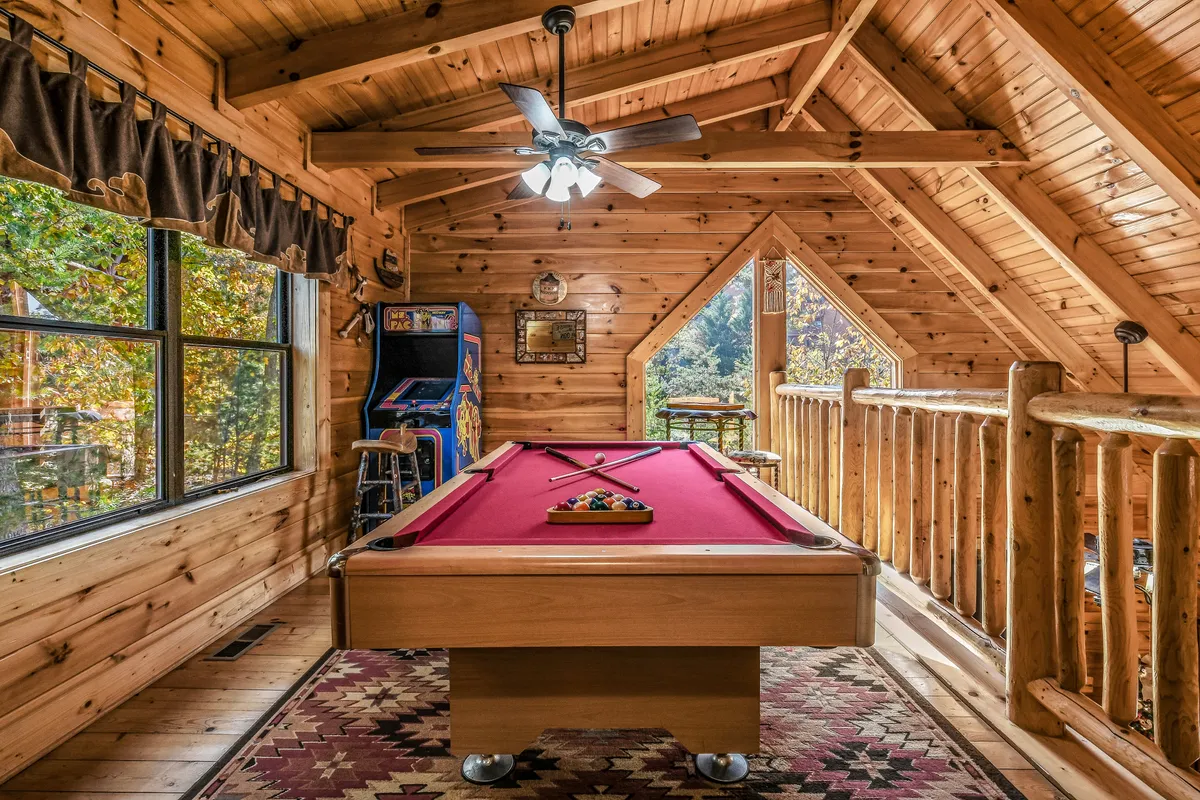 This inviting loft scene showcases a pool table positioned beneath a vaulted wooden ceiling, with expansive windows that offer views outside. An arcade game machine sits adjacent, blending classic fun with a warm, rustic atmosphere ideal for gatherings.