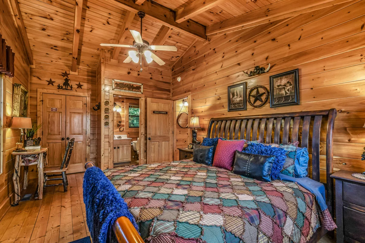 This image shows a cozy bedroom with a patchwork quilt, wooden walls, ceiling fan, decorative wall art, and wooden furniture. The room exudes a sense of warmth, privacy, and home-like comfort—ideal for restful nights or family stays.