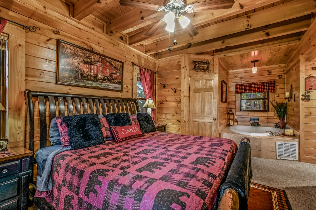 This image presents a cozy bedroom featuring a bed with a patterned blanket, decorative wall art, and a bathtub tucked into the corner. Warm wood paneling and a window that lets in natural light create a tranquil, rustic ambiance. The visible tub in the bedroom area hints at a private, indulgent bathing experience, aligning with the heart-shaped Jacuzzi tub mentioned in the section.