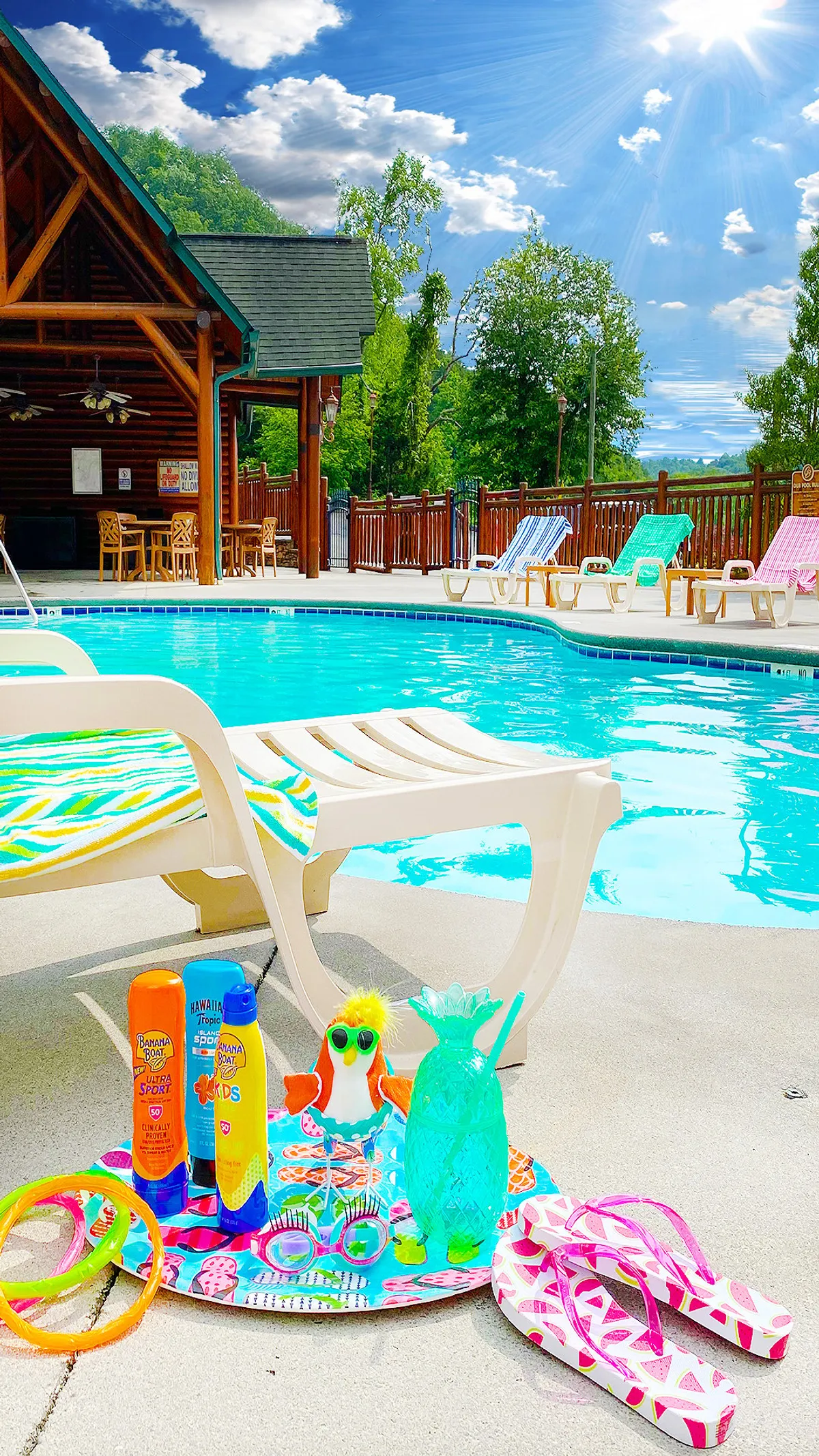 A vibrant outdoor scene with a large swimming pool surrounded by colorful pool floats, relaxing lounge chairs, and lush greenery. The cabin structure is visible in the background, inviting both energetic play and sun-soaked tranquility.