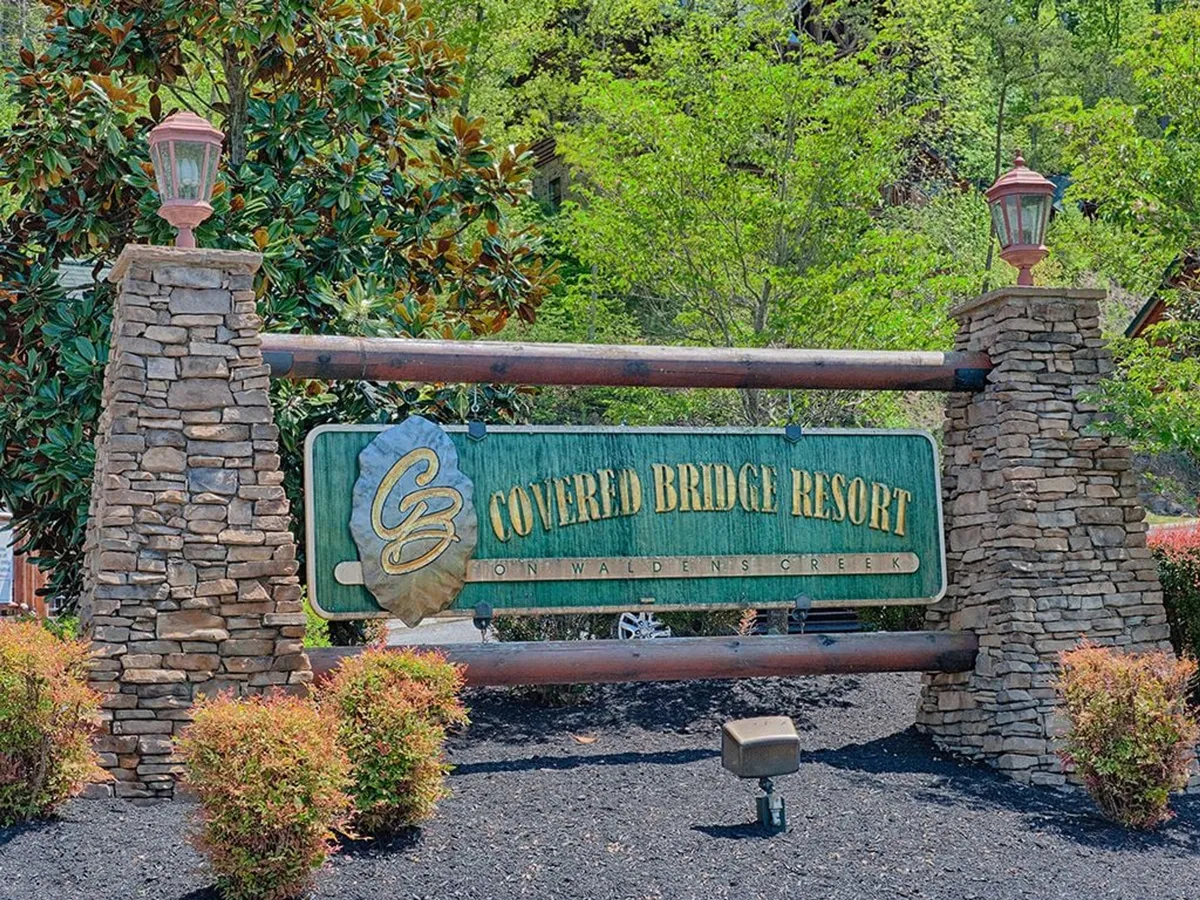 An inviting exterior shot showcasing a picturesque resort entrance with a stone sign, wooden beams, and lush greenery. Lamp posts and landscaping create a welcoming, communal feel, suggesting the start of a memorable family getaway.