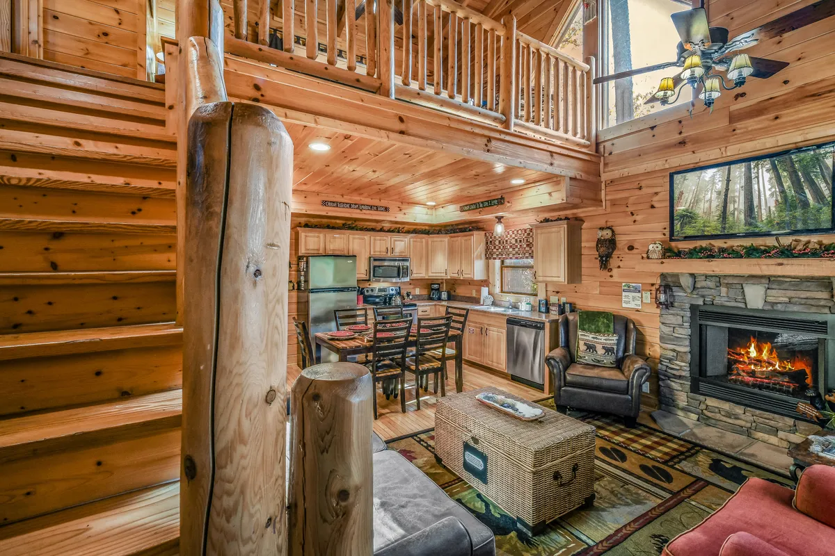 This image captures the heart of the open-concept living area with a view of the fireplace, staircase, kitchen, dining table, and a cozy seating area. Warm wood tones and rustic décor are enhanced by natural light from expansive windows.