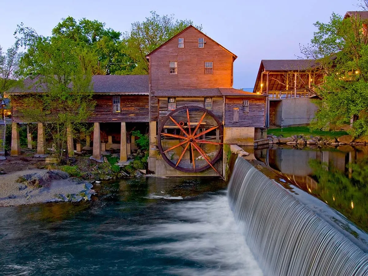 A picturesque exterior scene featuring a historic water wheel and waterfall beside a rustic wooden structure, nestled among lush trees. The tranquil setting offers a sense of place and connection to the natural attractions of the Smoky Mountains.