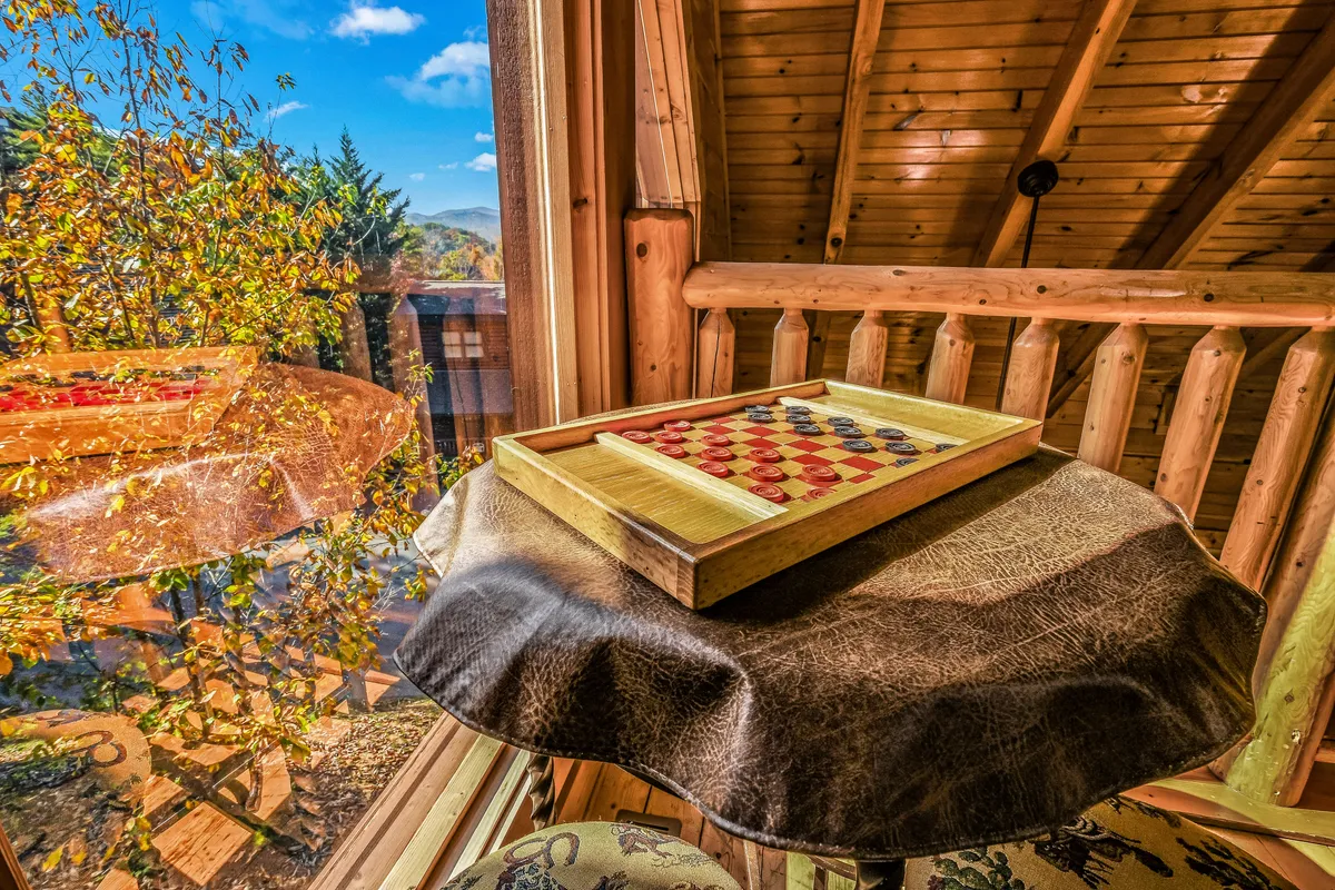 A rustic loft corner with a wooden checkerboard game set on a tray table, accented by exposed beams and expansive windows that reveal lush outdoor scenery. The setup invites casual play and conversation in a cozy, light-filled environment.