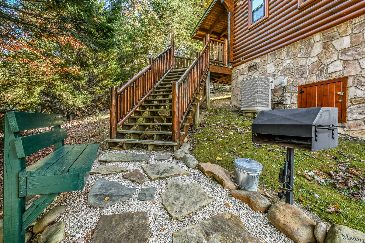 An exterior view highlighting a wooden staircase leading up to a porch, a green bench perfect for quiet moments, and a grill situated nearby. The stone pathway and surrounding greenery create a welcoming environment for outdoor relaxation and group barbecues.