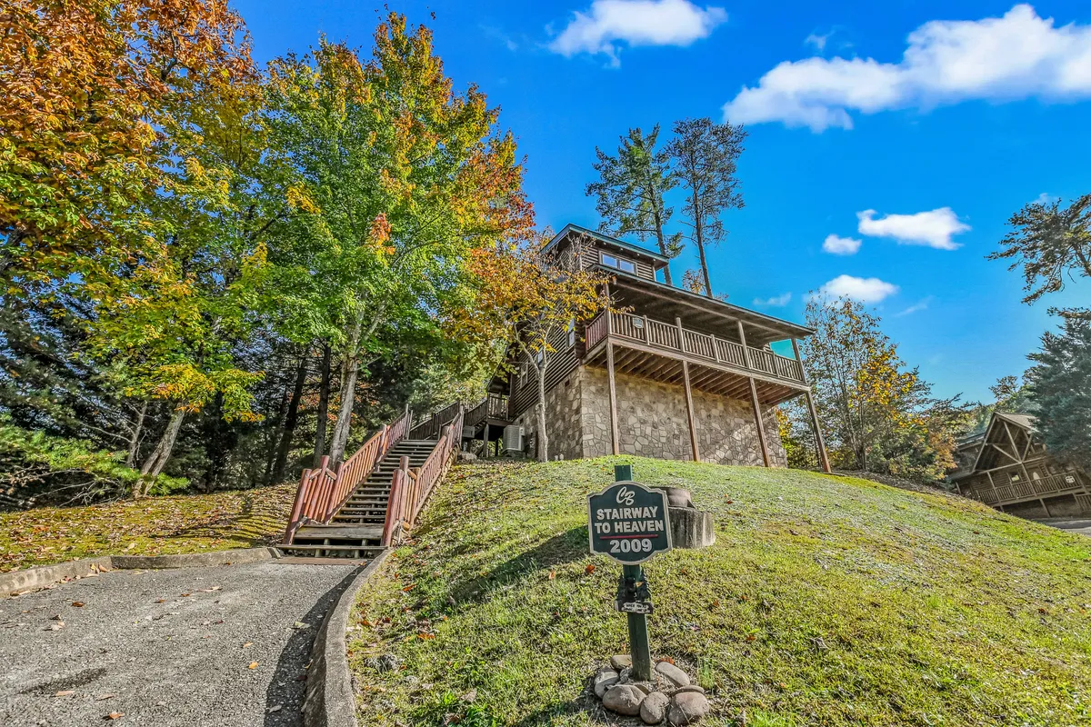 This image captures the stairway leading up to the cabin, surrounded by lush trees and featuring the welcoming 'STAIRWAY TO HEAVEN' sign. The scene is tranquil, with the home nestled amidst nature and a deck that hints at spaces for outdoor gatherings.