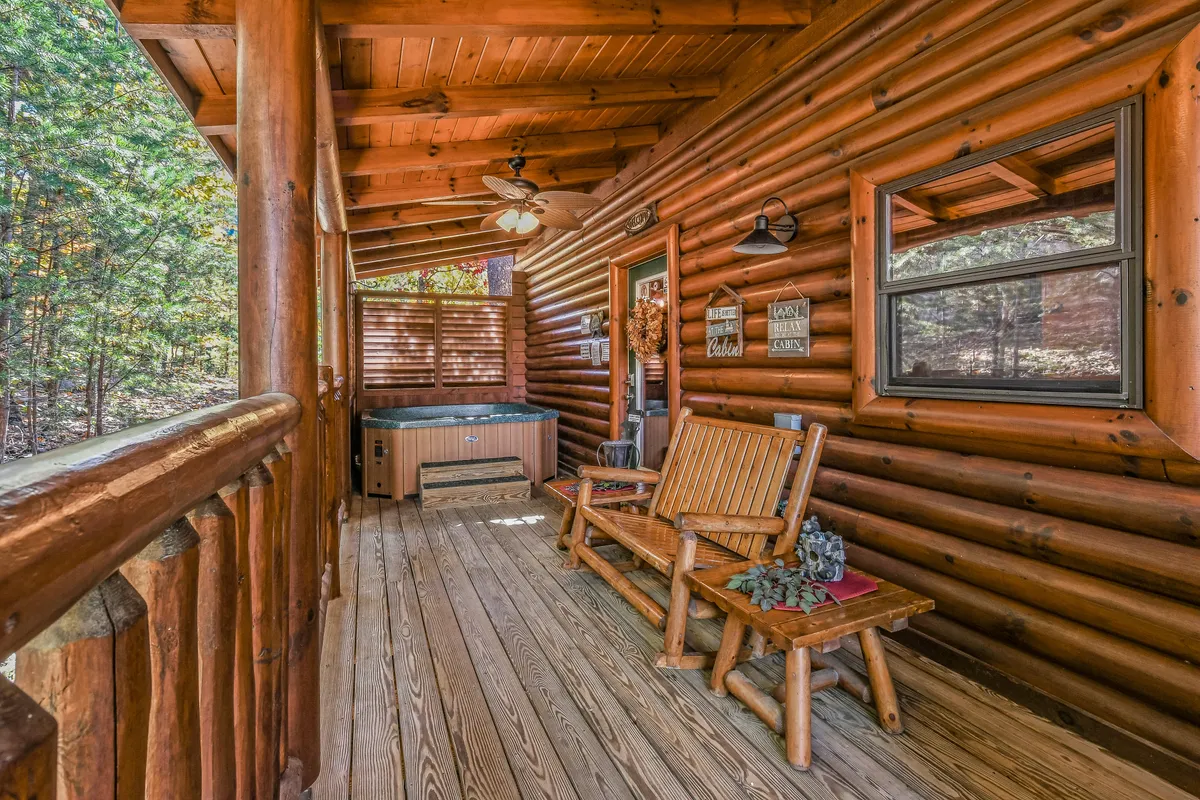 An exterior shot of a cozy log cabin deck features a bubbling hot tub, wooden benches, and rustic log walls, all set against a peaceful mountain backdrop. The setup encourages stargazing and relaxation while soaking in the hot tub.