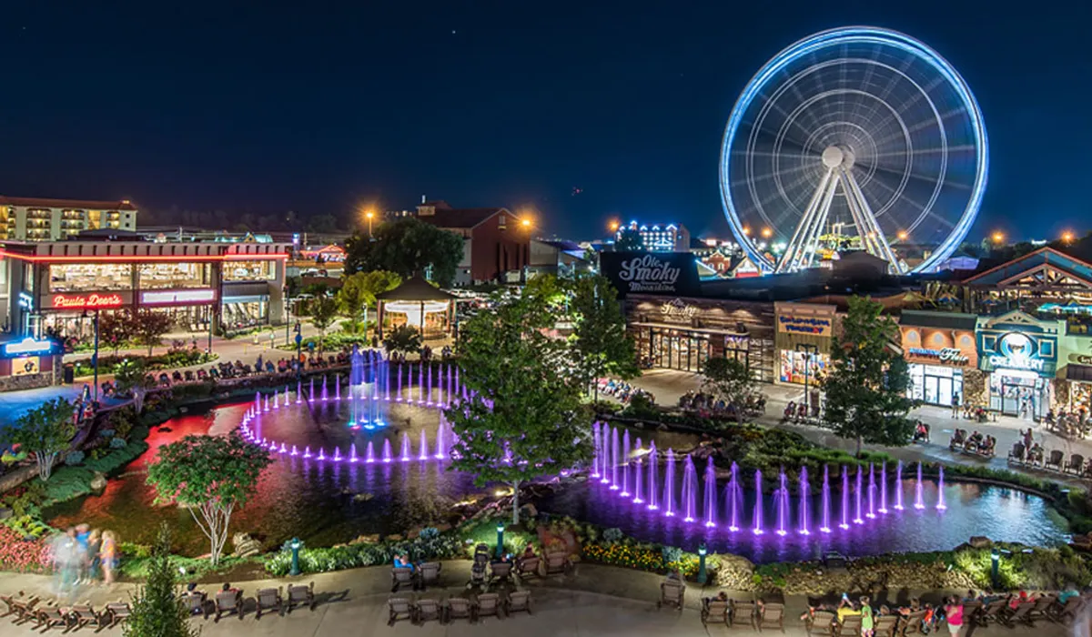 A lively nighttime scene of a vibrant shopping and entertainment area, featuring a Ferris wheel, a colorful fountain, illuminated pathways, and bustling people. The image captures the excitement and energy of local attractions and nightlife.
